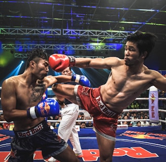 Two athletes are engaged in a dynamic kickboxing match inside a ring, surrounded by a cheering audience. One fighter, wearing red gloves and shorts, executes a high kick towards his opponent, who attempts to block the attack with his arm. The background features colorful lighting and promotional banners.