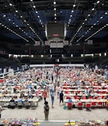 An indoor sports arena filled with rows of tables, each occupied by people engaged in competitive activities. The ceiling is high, with numerous lights illuminating the large space. The audience seating on either side is empty, and there are flags hanging from the upper level.