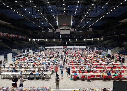 An indoor sports arena filled with rows of tables, each occupied by people engaged in competitive activities. The ceiling is high, with numerous lights illuminating the large space. The audience seating on either side is empty, and there are flags hanging from the upper level.