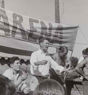 A group of men, some shirtless, are gathered around a platform or stage. A man in a white shirt appears to be speaking or gesturing to a crowd. A large sign with the word 'ARENA' is prominently displayed in the background. The scene seems to be set outdoors on a sunny day.
