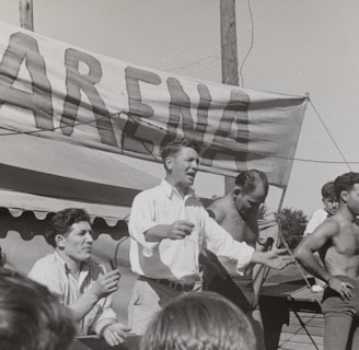 A group of men, some shirtless, are gathered around a platform or stage. A man in a white shirt appears to be speaking or gesturing to a crowd. A large sign with the word 'ARENA' is prominently displayed in the background. The scene seems to be set outdoors on a sunny day.