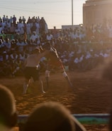 A vibrant wrestling match taking place outdoors, surrounded by a large crowd of spectators. The atmosphere is charged with excitement as people stand and sit around the dirt arena, intently watching the competitors. The sun is setting, casting a warm glow over the scene.