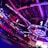 An indoor arena with vibrant purple and blue lighting, featuring large numbered screens suspended from the ceiling. The venue has a high ceiling with intricate metal truss structures and hanging lights, creating a futuristic and energetic atmosphere.