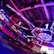 An indoor arena with vibrant purple and blue lighting, featuring large numbered screens suspended from the ceiling. The venue has a high ceiling with intricate metal truss structures and hanging lights, creating a futuristic and energetic atmosphere.