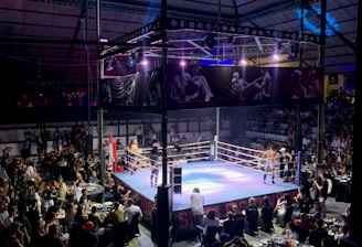 A crowded indoor arena with a boxing ring at the center, surrounded by an audience seated at tables. The scene is illuminated by overhead lights, casting a vibrant glow over the ring where two fighters are preparing for a match. Spectators are engaged, capturing the event with cameras and enjoying the lively atmosphere.