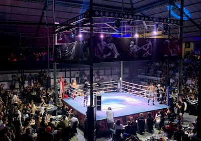 A crowded indoor arena with a boxing ring at the center, surrounded by an audience seated at tables. The scene is illuminated by overhead lights, casting a vibrant glow over the ring where two fighters are preparing for a match. Spectators are engaged, capturing the event with cameras and enjoying the lively atmosphere.