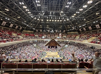 A large indoor arena packed with spectators seated in tiers around a central platform. The platform features traditional Japanese decor, indicating it may be used for sumo wrestling. The atmosphere is bustling, with people filling the seats and many spectators visible in the foreground.