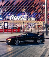 A sleek black sports car is parked in front of an illuminated arena entrance, with a large sign bearing the name of a famous beverage brand. The building features a modern, geometric facade with bright red and blue lights, creating a vibrant and lively atmosphere.